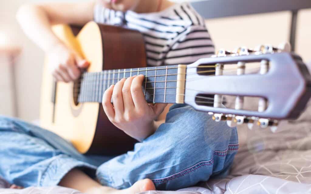 Young girl sitting cross legged on a bed playing an acoustic guitar