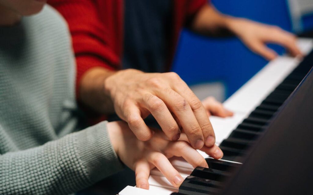 Fingers of a boy sitting next to his piano teacher helping him play the piano