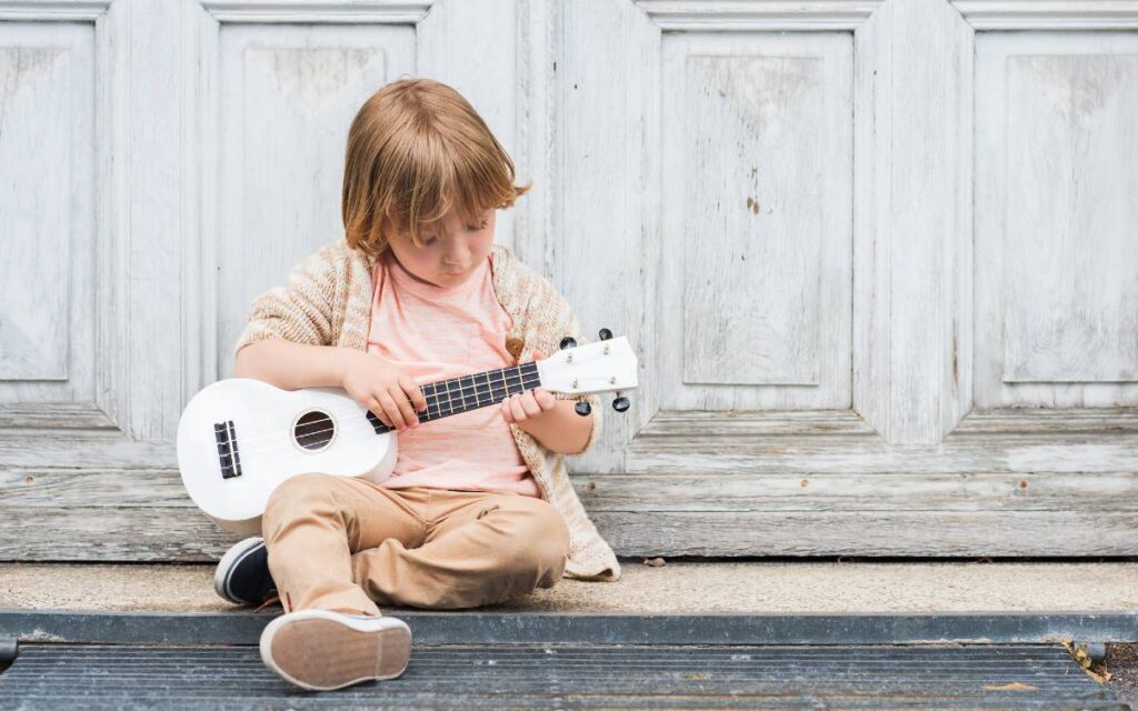 Little boy sitting against wooden door playing ukulele