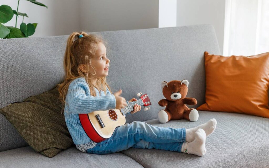 Little girl sitting on a sofa next to her stuffed toy, playing ukulele