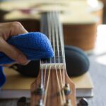 A man's hand cleaning acoustic guitar strings using a blue cloth